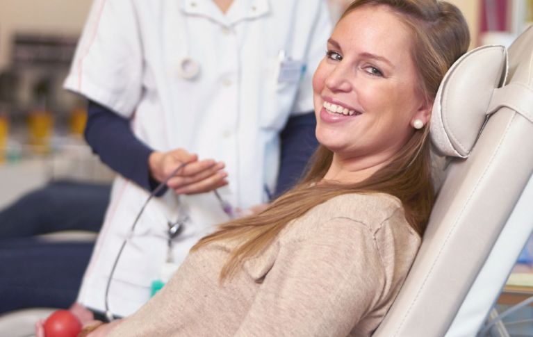 Eine junge Frau lächelt in die Kamera und spendet Blut. Im Hintergrund medizinisches Personal mit Schlauch in der Hand. 